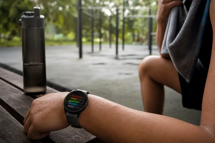 Polso con orologio sportivo, borraccia e atleta in pausa all’ombra.