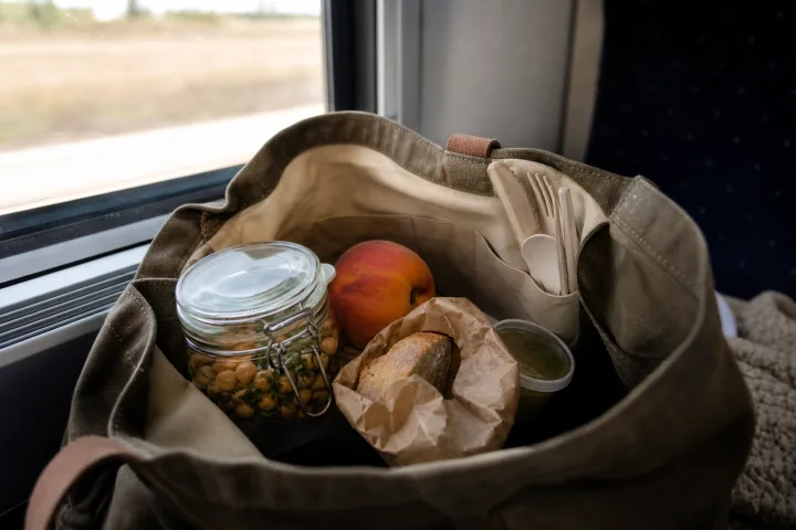Pranzo al sacco in una borsa da viaggio con frutta intera, pane avvolto e posate riutilizzabili.