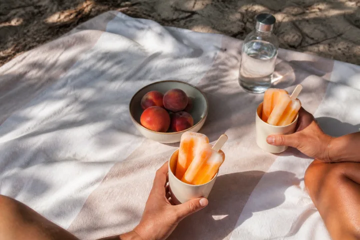 Due mani tengono ghiaccioli alla frutta su un telo da spiaggia con pesche e acqua accanto.